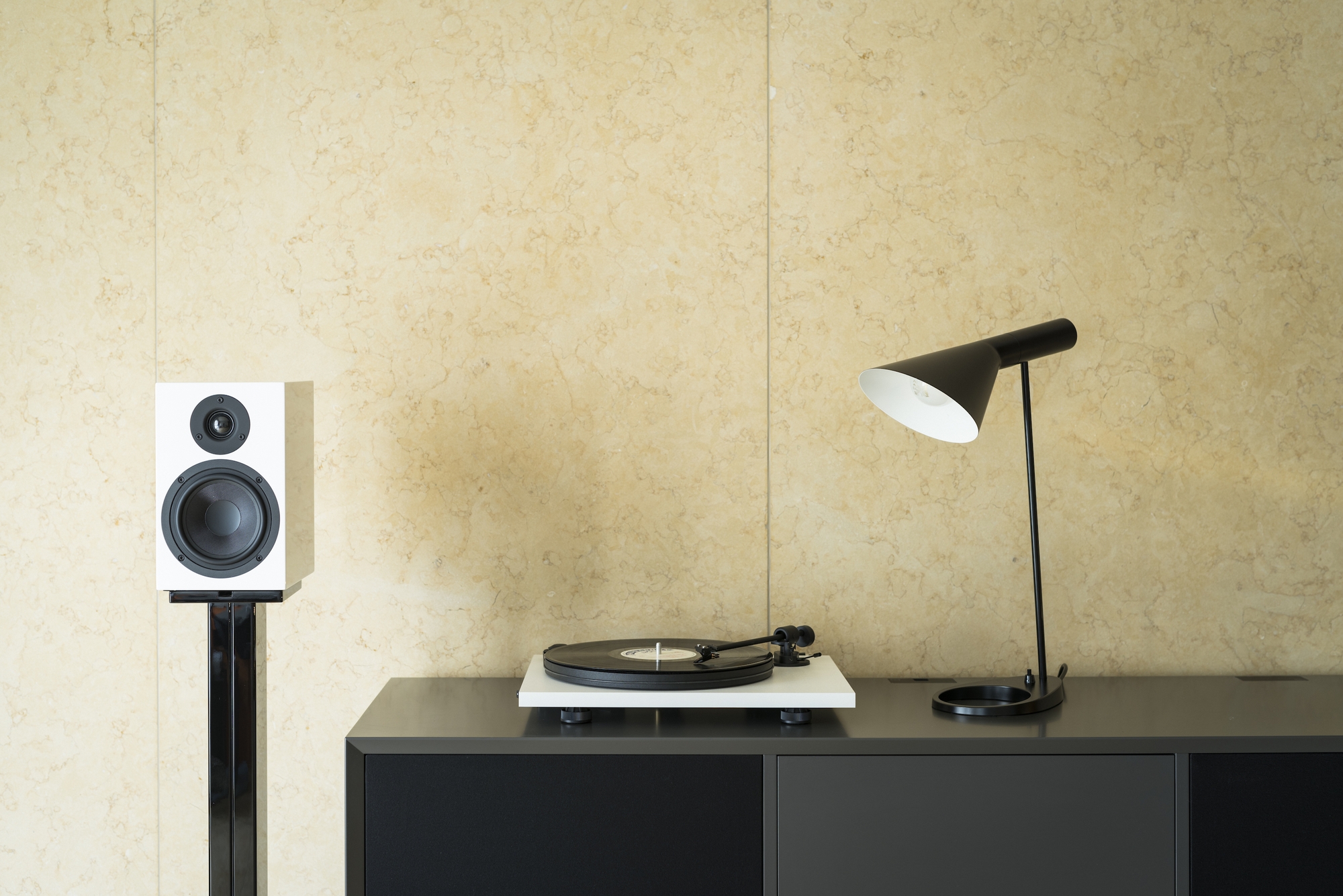 Pro-Ject Primary E Turntable With Om Cartridge White turntable on a black cabinet, white speaker on a stand, and a black floor lamp with white shade.