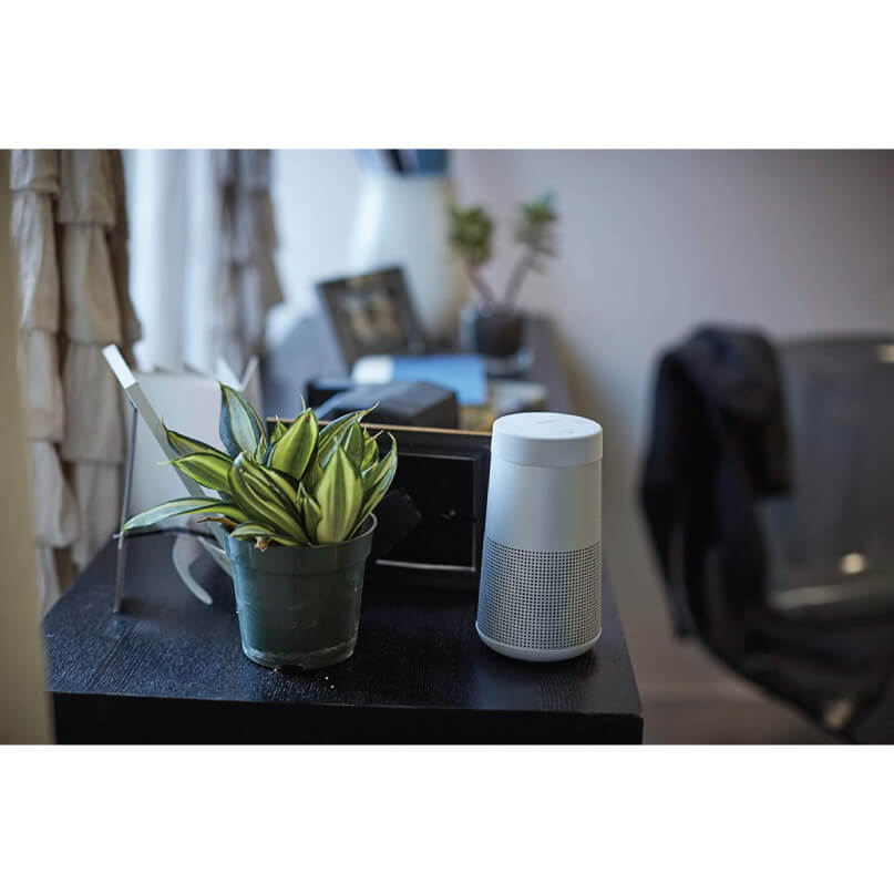 Bose Soundlink Revolve+ Bluetooth Speaker Amazon echo fourth generation smart speaker, white cylindrical with perforated mesh, on a dark desk beside a potted plant.