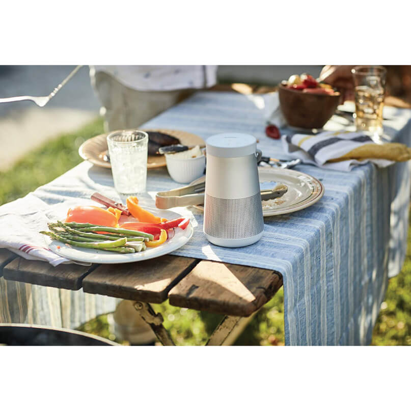 Bose Soundlink Revolve+ Bluetooth Speaker Portable speaker with a silver body and perforated lower grille on a blue striped picnic table outdoors.