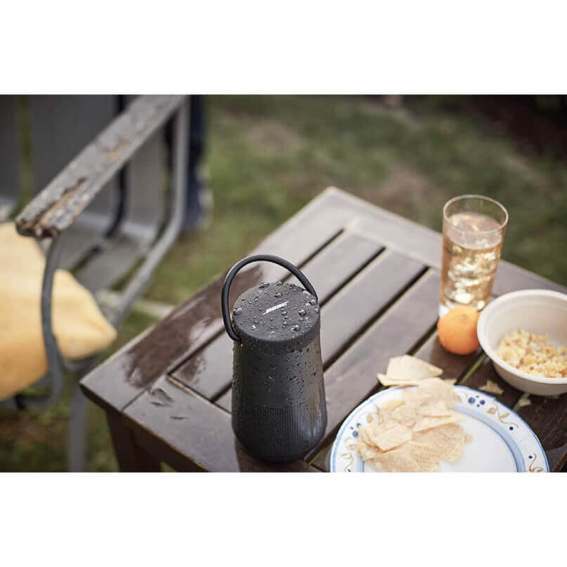 Bose Soundlink Revolve+ Bluetooth Speaker Black cylindrical portable bluetooth speaker with a loop handle, wet surface, on a wooden outdoor table beside snacks.
