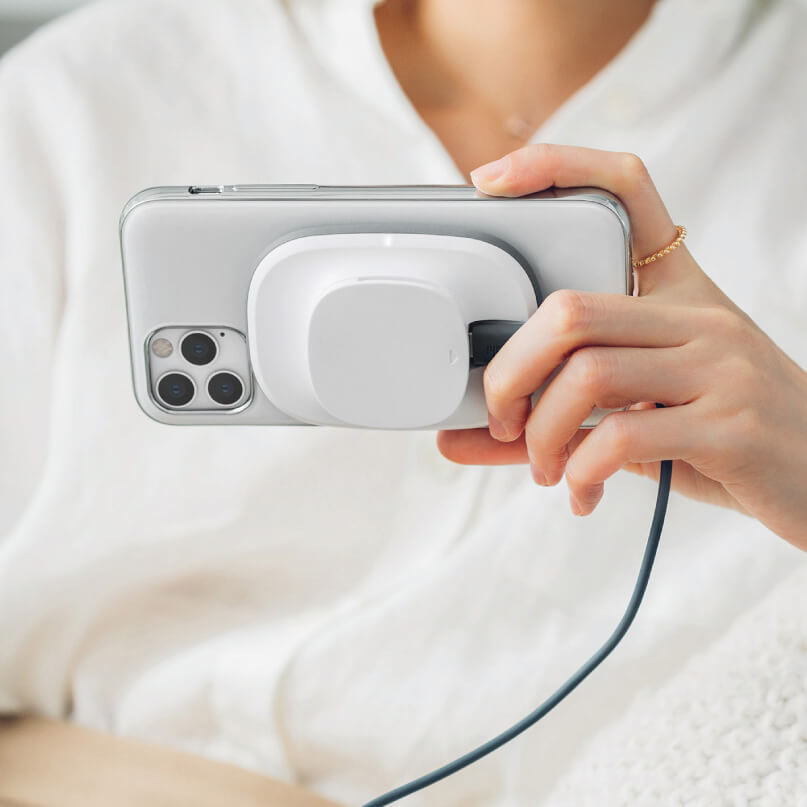 Moshi Snapto Magnetic Wireless Charger W/ Wall Mount Kit (Grey) A person holds a smartphone with a circular white wireless charging puck attached to its back.