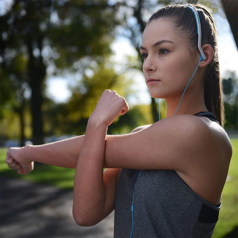 Koss Kph14 On Ear Headphones Young woman in a gray athletic top wearing blue wired earphones outdoors, stretching her arms.