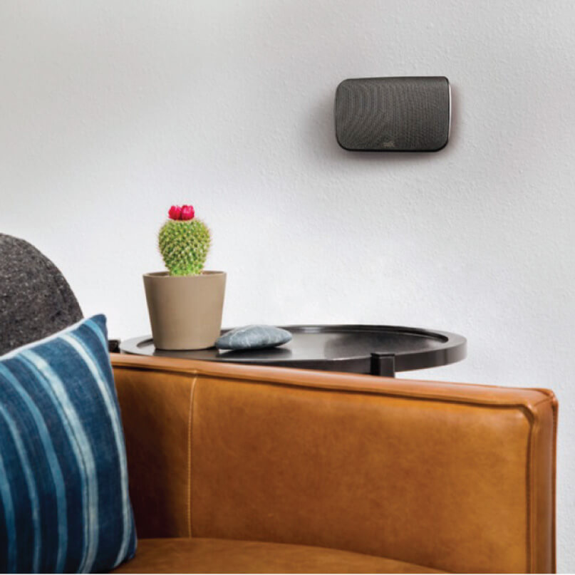 Polk Audio Sr1 Wireless Surround Speakers For Magnifi Max Black wall-mounted wireless speaker with rounded rectangular design above a round black table and potted cactus.