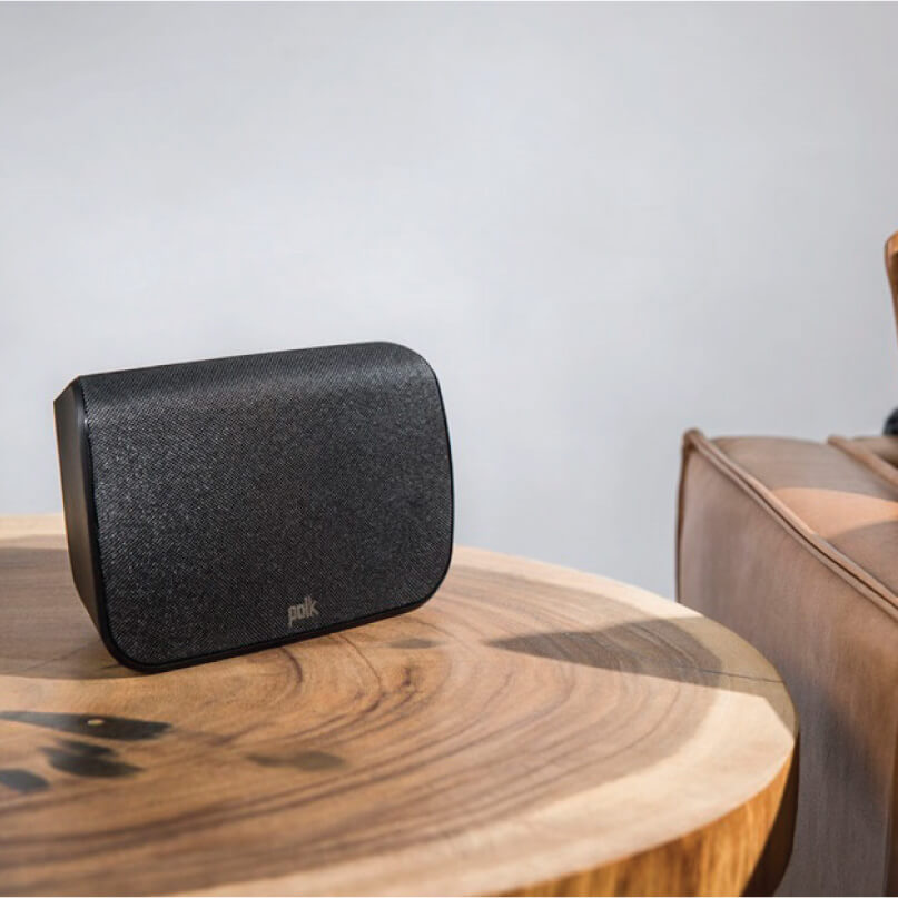 Polk Audio Sr1 Wireless Surround Speakers For Magnifi Max Polk portable wireless speaker with black fabric grille and rounded rectangular body on a round wooden table.