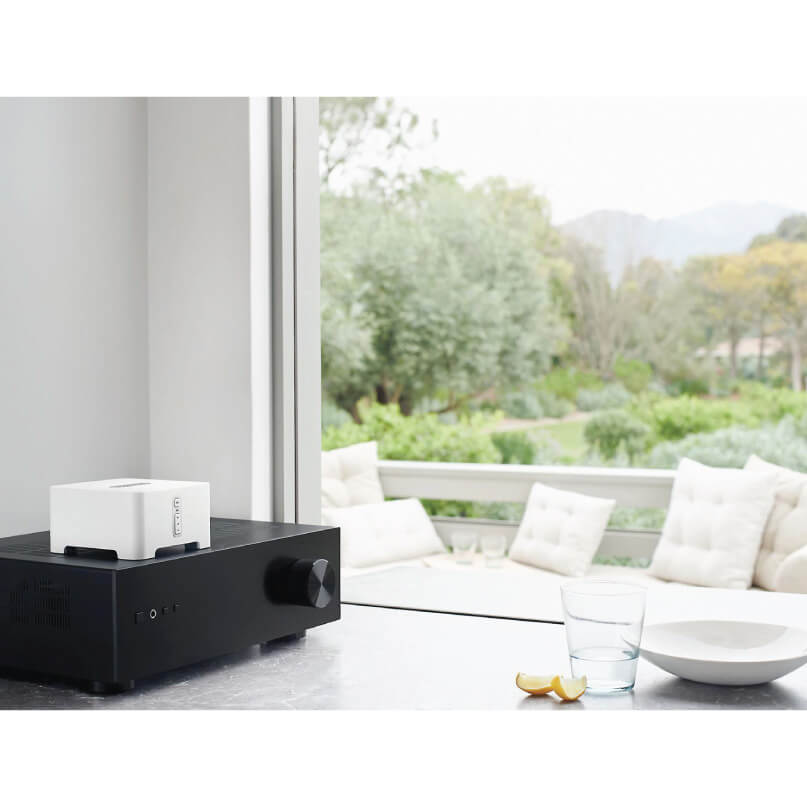 Sonos Connect - Turn Home Theatre Into Music Streaming System Black stereo amplifier with a white rectangular top unit and a large front knob, on a counter by a window.