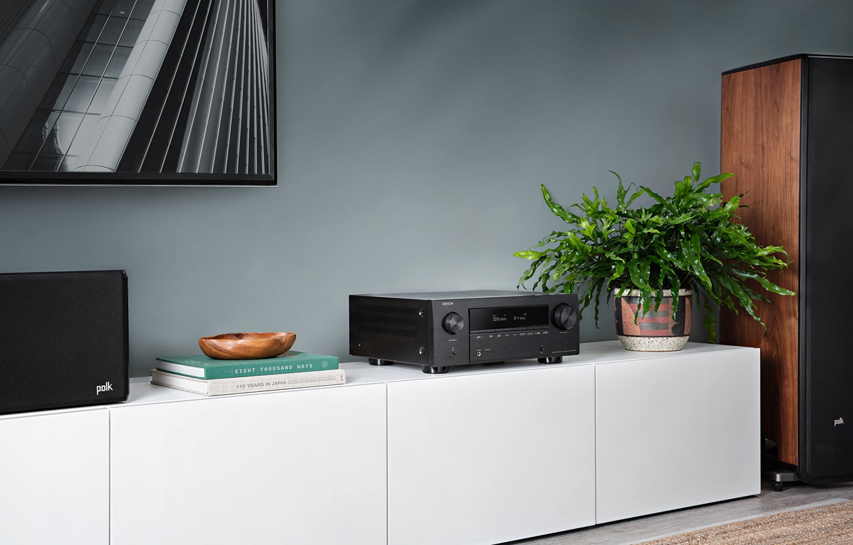 Denon Avc-X3700H Denon stereo receiver on a white cabinet, flanked by polk speakers, with a potted plant and books nearby.