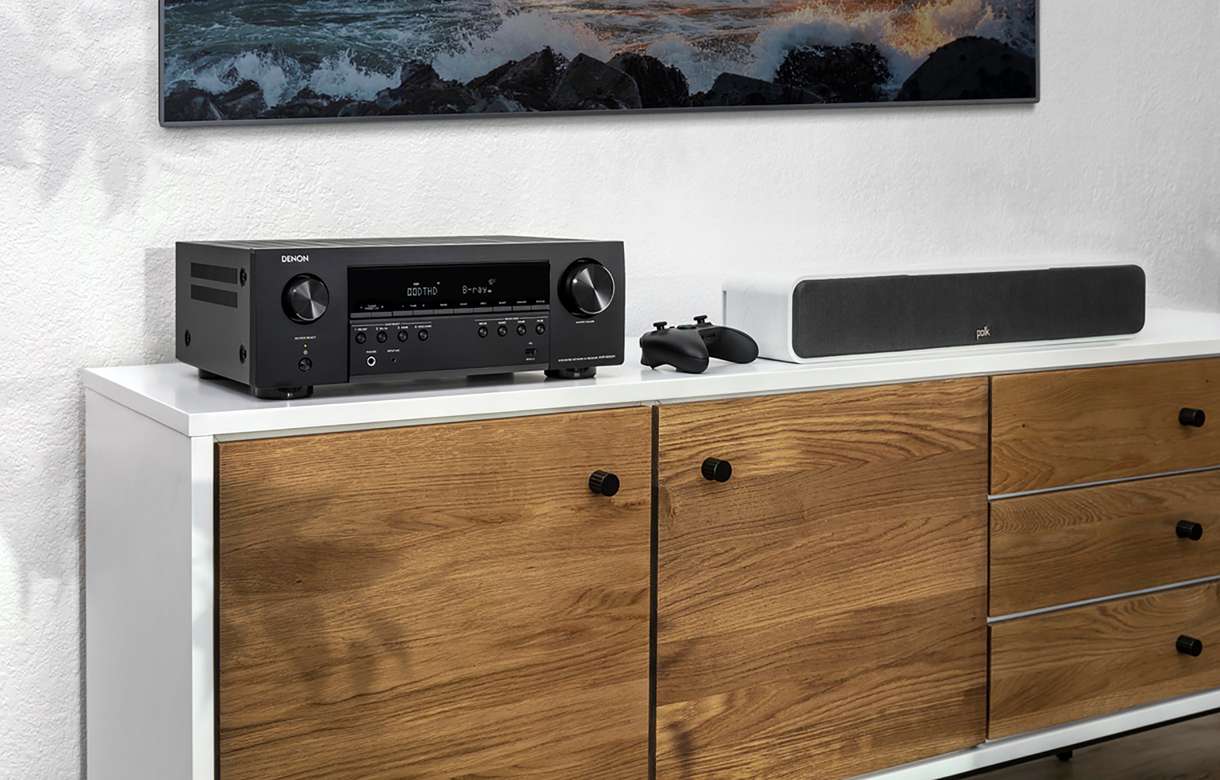 Denon Avr-S660H Denon av receiver with front-panel controls on a white cabinet, beside a polk soundbar and a game controller.