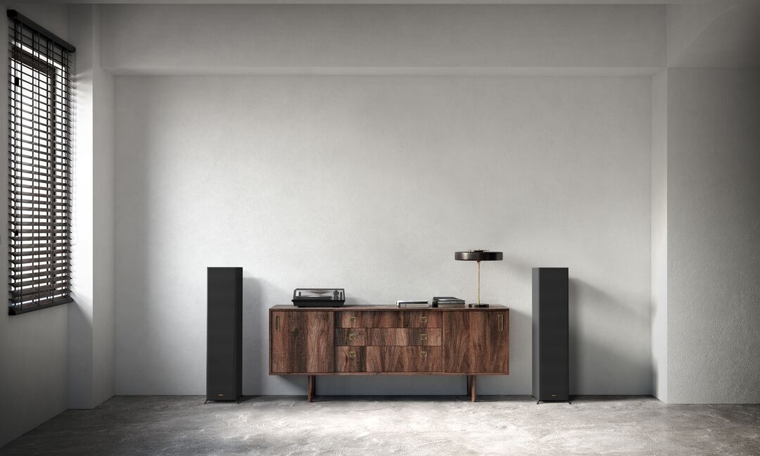 Klipsch Rp-8060Fa Ii Dolby Atmos Floorstanding Speakers Walnut sideboard flanked by two tall black floor-standing speakers, with a turntable and lamp on top in a minimalist room.