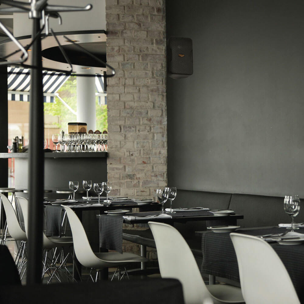 Klipsch Cp-6T - Sold As Pair Modern restaurant dining area with brick pillar, dark tables, and white chairs set with wine glasses.