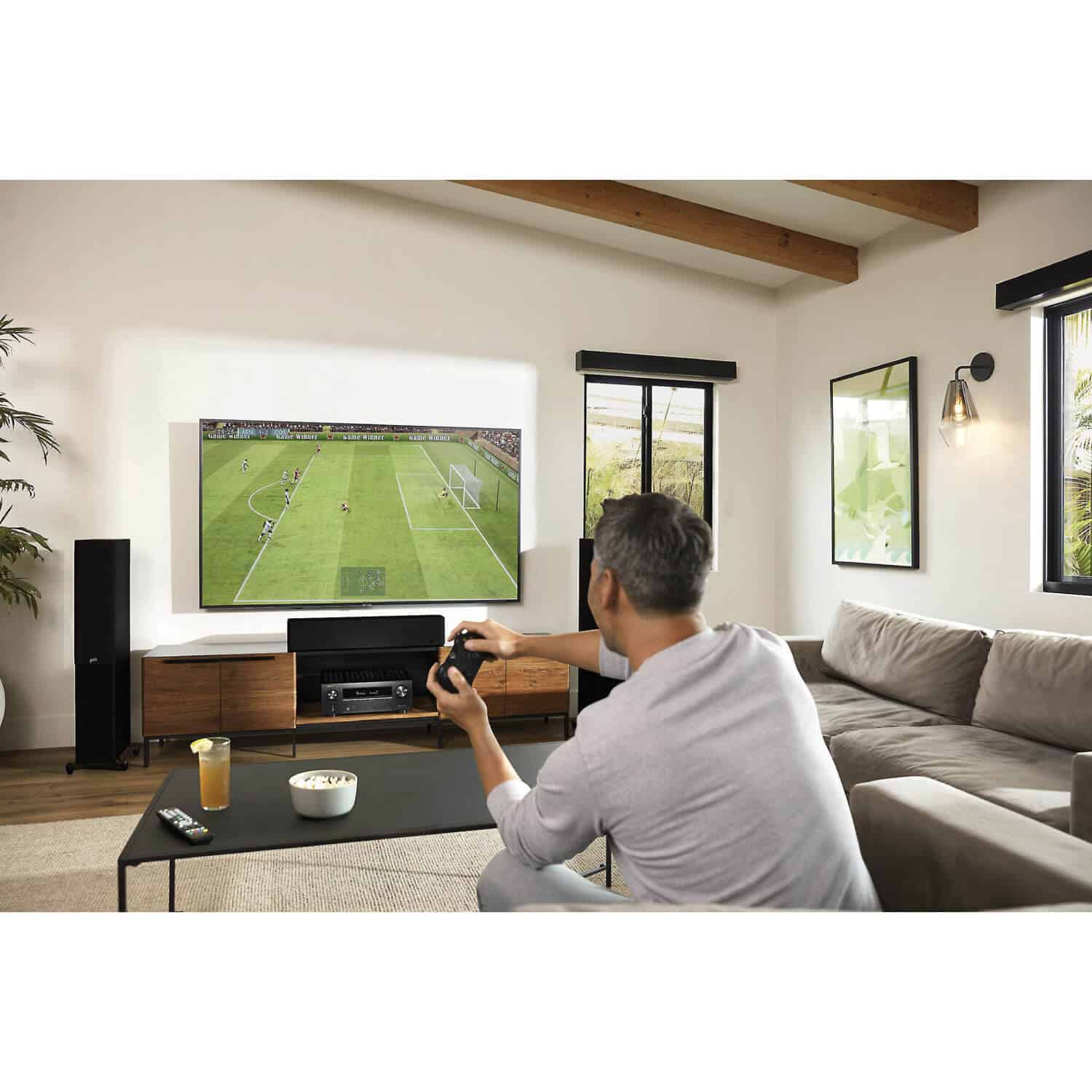 Denon Avr-X2800H 7.2Ch 8K Av Receiver Man playing a soccer video game on a large flat-screen tv in a modern living room.