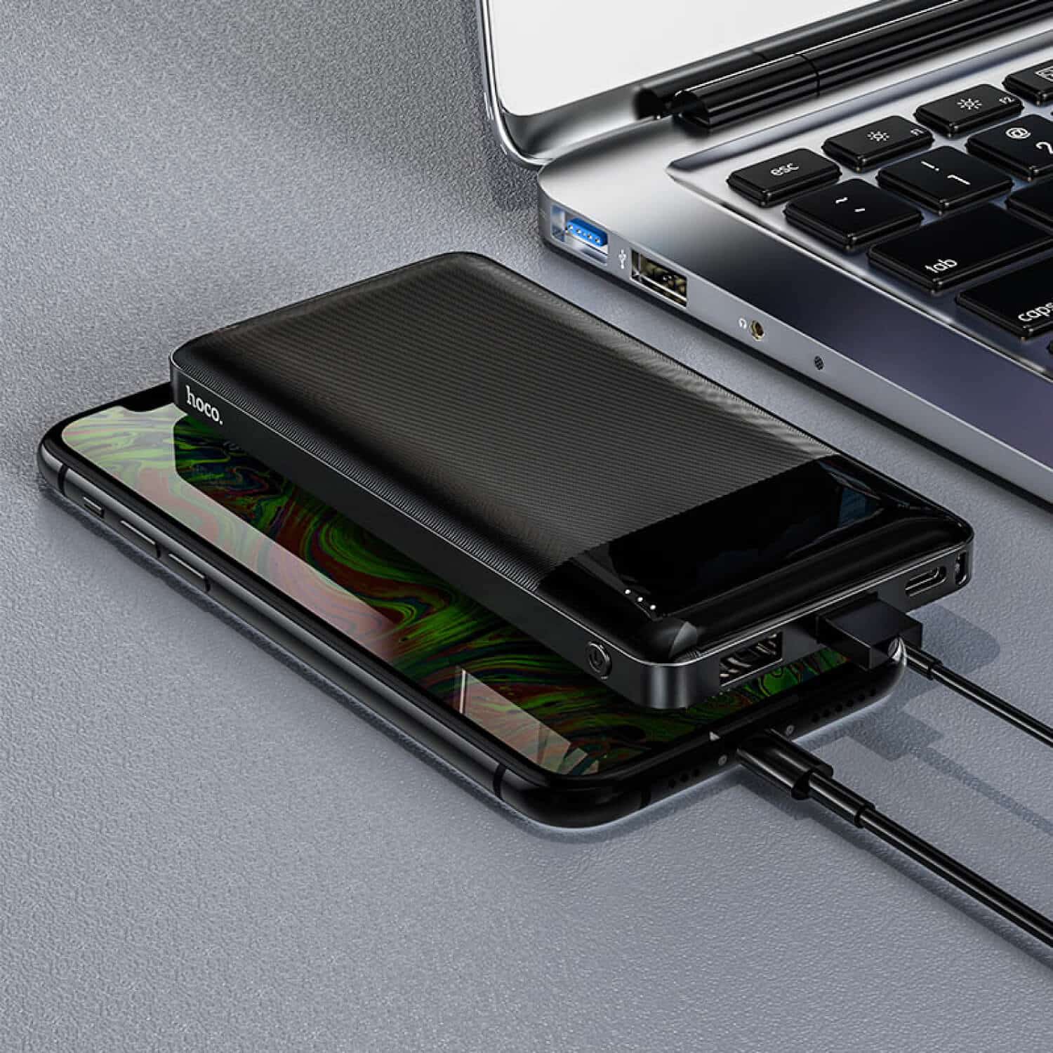 Hoco Power Bank “J72 Easy Travel” 10000Mah Hoco power bank with a textured black surface beside a smartphone and a silver laptop with usb ports.