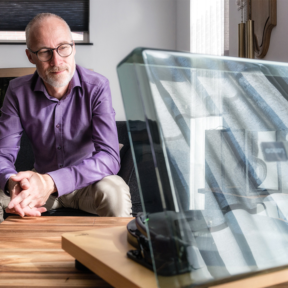 Lenco Turntable With Mmc Cartridge - Wood Older man with glasses in a purple shirt sits at a wooden table, facing a large curved glass computer monitor.