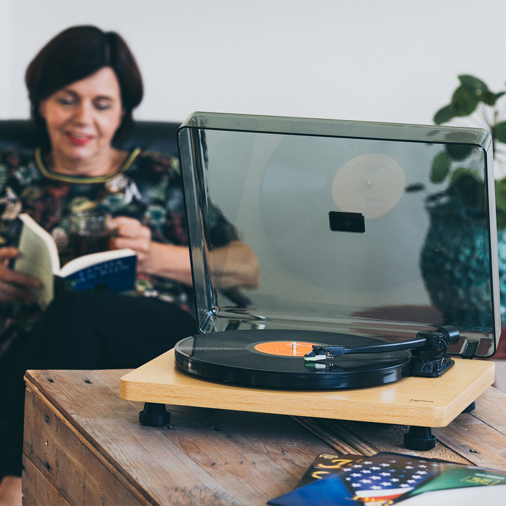 Lenco Turntable With Mmc Cartridge - Wood Turntable with clear lid on a wooden base, spinning orange-labeled vinyl while a woman reads in the background.
