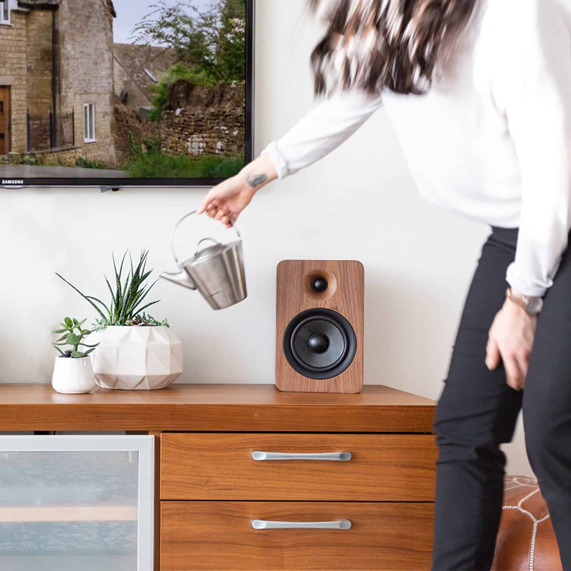 Kanto Yu6 200W Powered Bookshelf Speakers With Bluetooth® And Phono Preamp - Pair, Walnut Samsung flat-screen tv displays outdoor scene; wooden speaker, potted plants, and a person holding a metal kettle nearby.