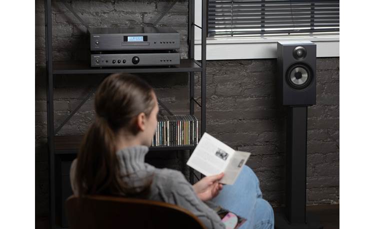 Rotel Cd11Mkii Cd Player Woman seated and reading a magazine beside a black stereo system on a metal shelf with a floor-standing speaker.