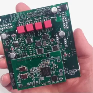 Handheld green printed circuit board (PCB) with pink rectangular capacitors and multiple ICs, plus a secondary module.