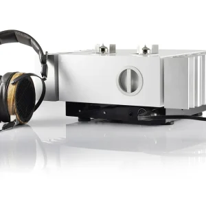 Silver headphone amplifier with large side heatsink fins and circular front dial, paired with wooden over-ear headphones.