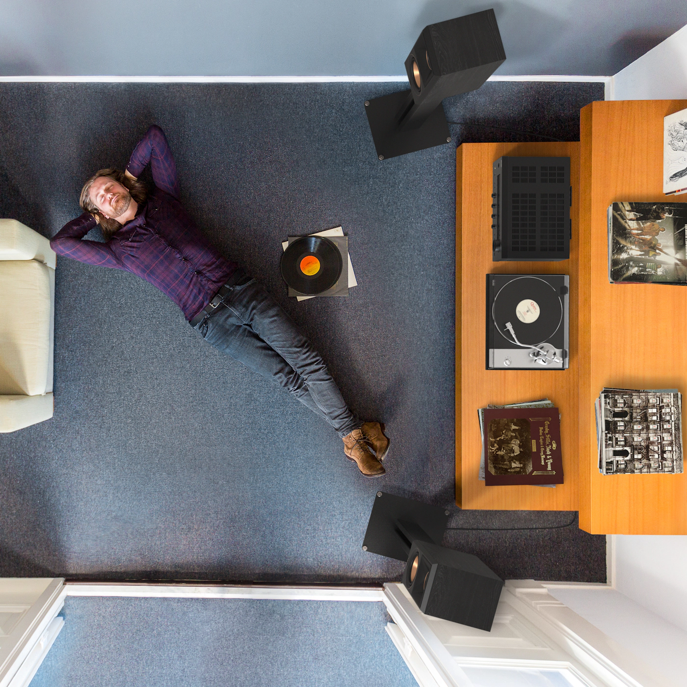 Man lying on blue carpet beside a wooden desk with a turntable, vinyl records, and black speakers