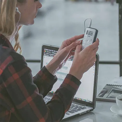 Sangean Dt-140 Am/Fm Pocket Receiver Person holds a small handheld digital device with a display and buttons beside a laptop.