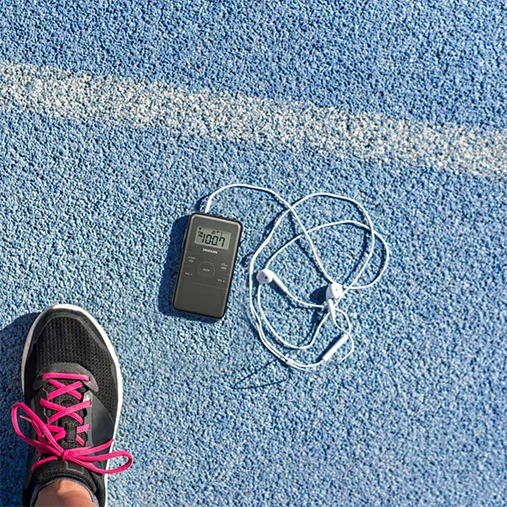 Sangean Dt-140 Am/Fm Pocket Receiver Black digital stopwatch with display and tangled white earbuds on a blue running track beside a sneaker.