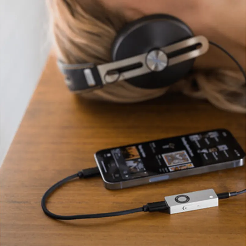 Audioengine Dac3 Portable Headphone Amplifier &Amp; Dac Over-ear headphones with a metal headband connected to a small silver portable dac/amp and a smartphone.