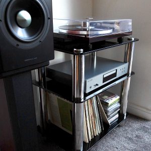 Silcron Steelseries 3 Shelf Hi-Fi Rack – Black Glass Turntable on a chrome glass shelf unit with a stereo receiver and vinyl records, beside a large speaker.