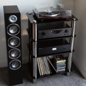 Silcron Steelseries 4 Shelf Hi-Fi Rack – Black Glass Tall black floor-standing speaker beside a glass hi-fi rack with a turntable on top and vinyl records below.