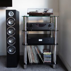 Silcron Steelseries 4 Shelf Hi-Fi Rack – Black Glass Black floor-standing speaker beside a glass audio equipment rack holding a turntable on top with an amplifier and vinyl records.