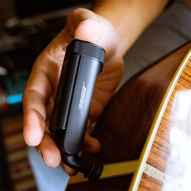 Bose Wireless Instrument Transmitter Bose clip-on microphone held in hand beside an acoustic guitar.
