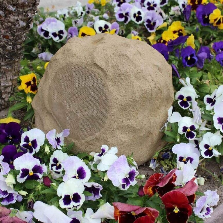Tgd Audio Ors-602G 6.5″ Outdoor Rock Speaker (Granite) Rock-shaped garden speaker with a circular mesh grill, nestled among colorful pansies.