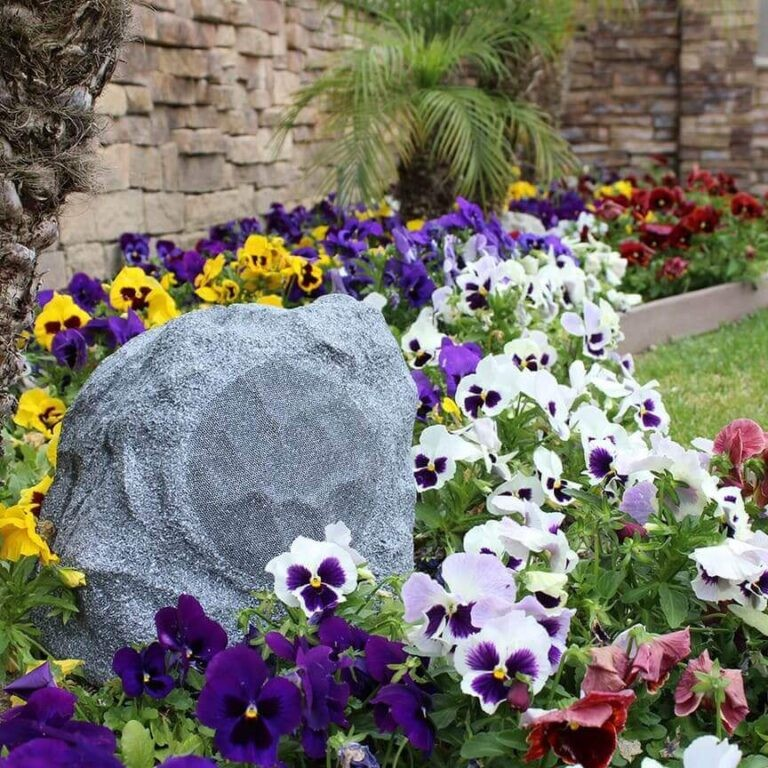 Tgd Audio Ors-602G 6.5″ Outdoor Rock Speaker (Granite) Decorative gray garden rock among colorful pansies beside a stone wall.