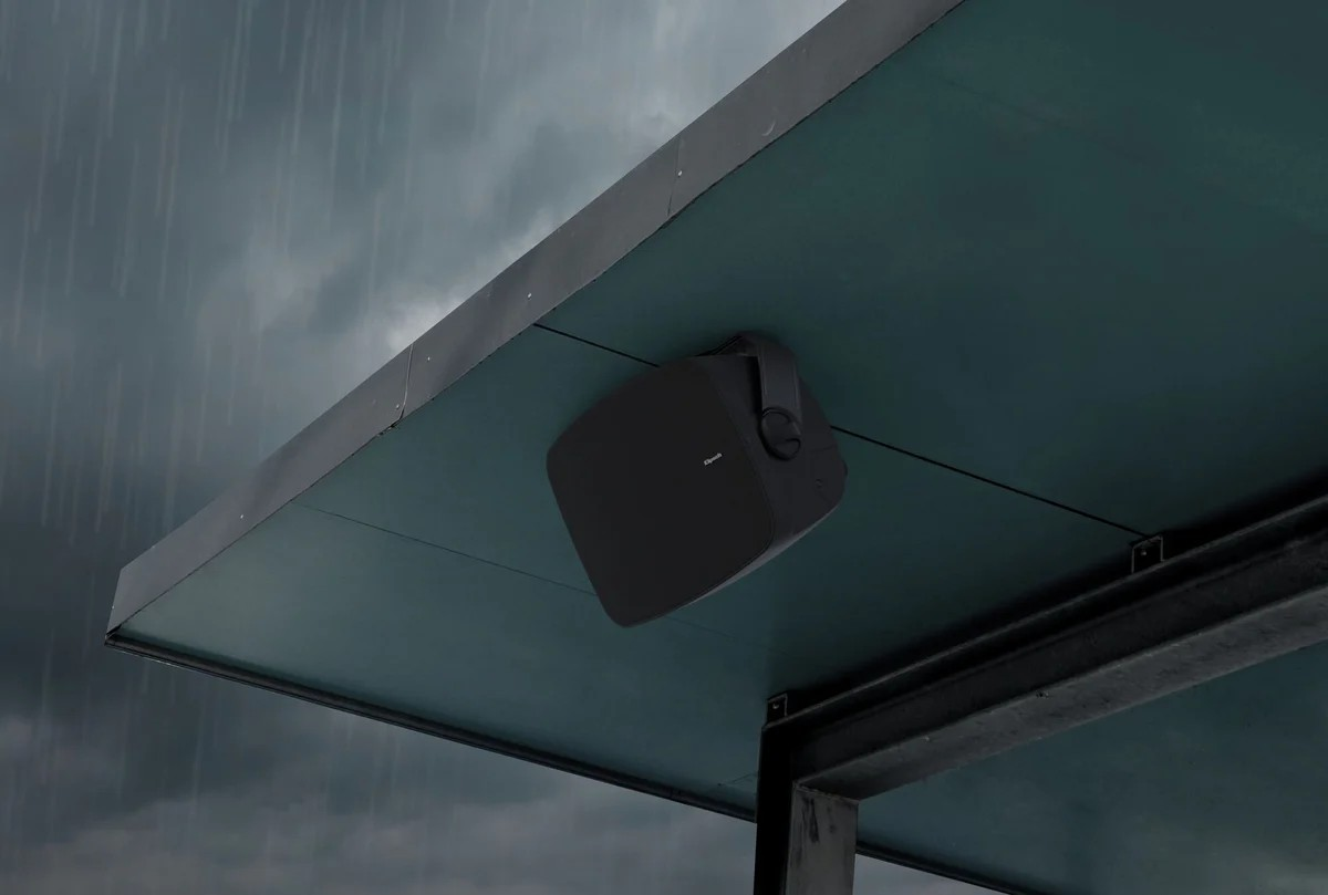 Klipsch Rsm-525 Bose outdoor wall-mounted speaker in black, square with rounded edges, under a metal canopy in rain.