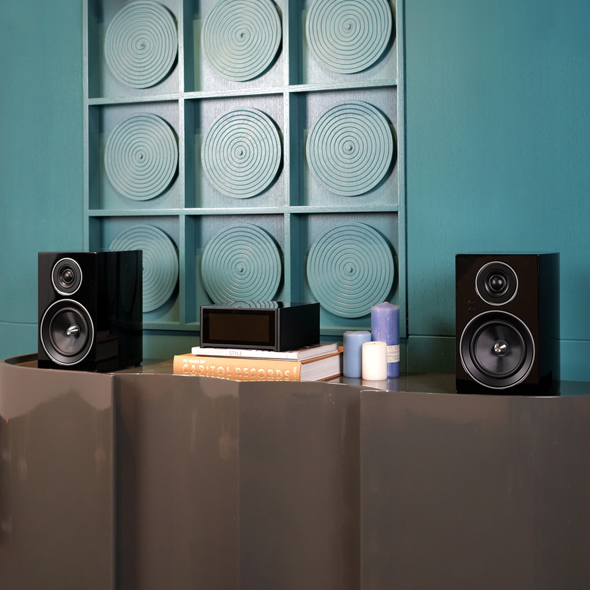 Jamo C707 Pa Active Bookshelf Speaker Two black bookshelf speakers flank a central stereo unit on teal cabinet.
