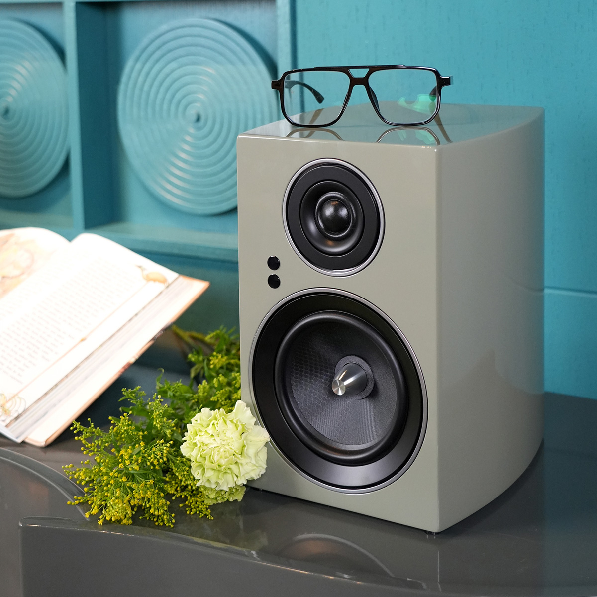 Jamo C707 Pa Active Bookshelf Speaker Gray bookshelf speaker with glasses on top, turquoise background.