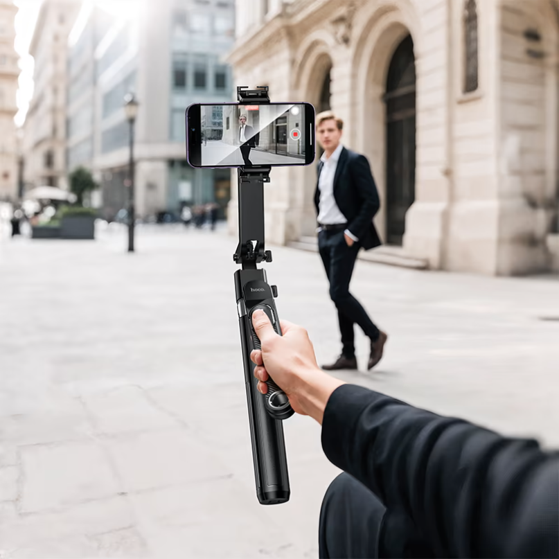 Hoco. Smart Holder “K32 Soporte” Quadropod Face Tracking A person is using the hoco. Smart holder “k32 soporte” quadropod face tracking with their smartphone to record a video on a city street, while a man walks by in the background.