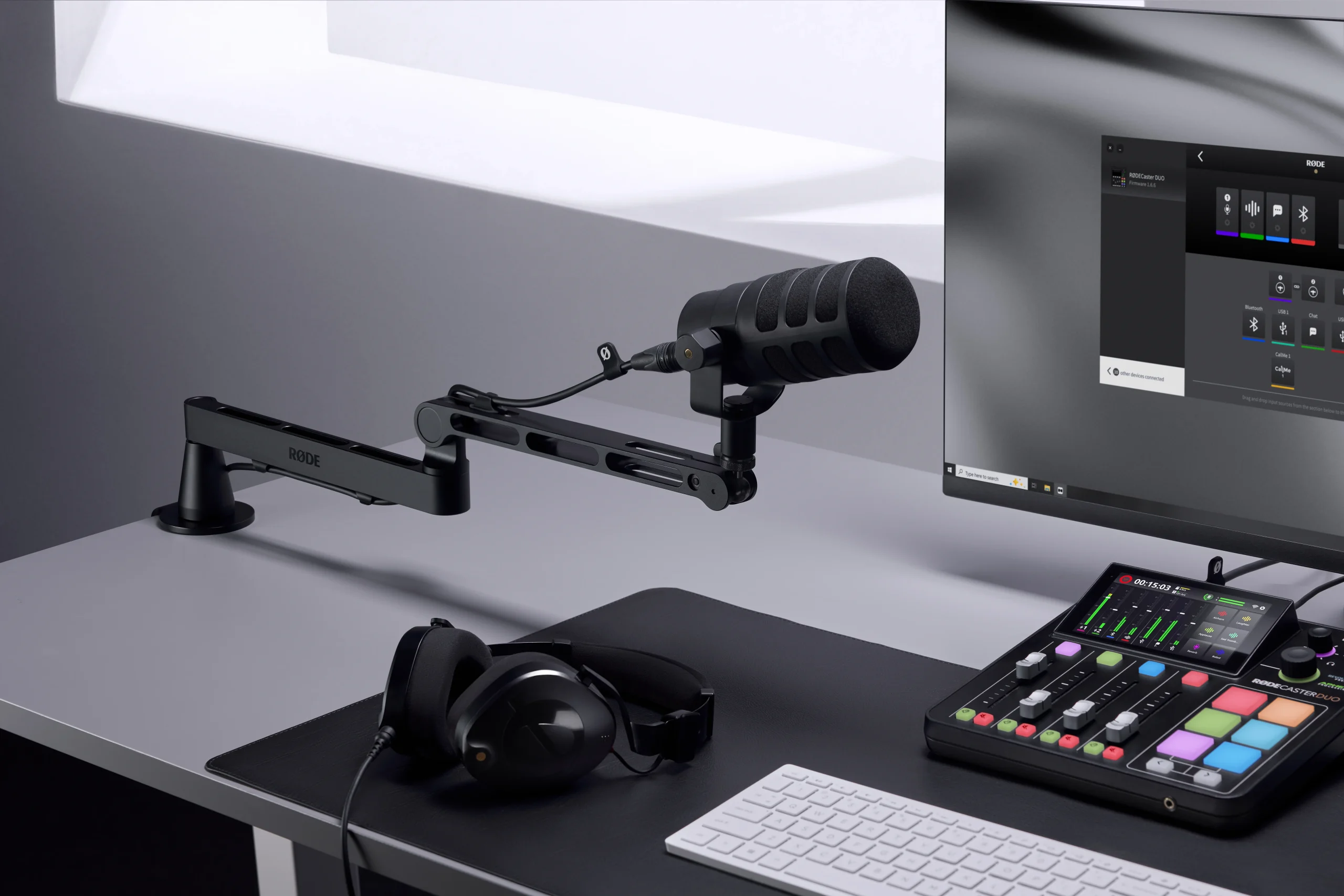 A black microphone mounted on a rode lpa1 low-profile studio arm, alongside headphones, a mixer, a keyboard, and a computer monitor arranged on a neatly organized desk.